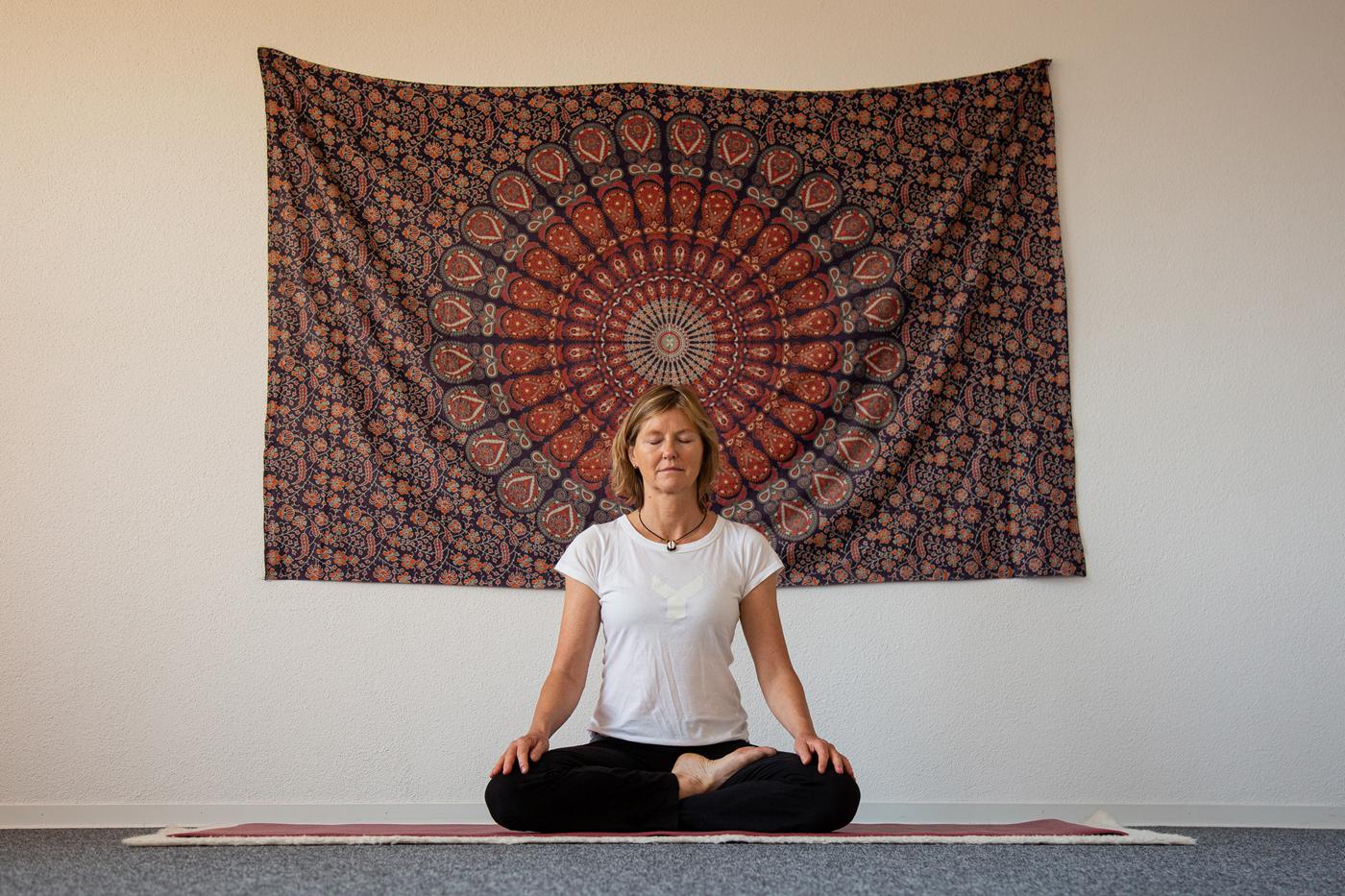 Jutta Thies sitzt in Meditationshaltung vor einem Mandala-Wandtuch im Yogazentrum
